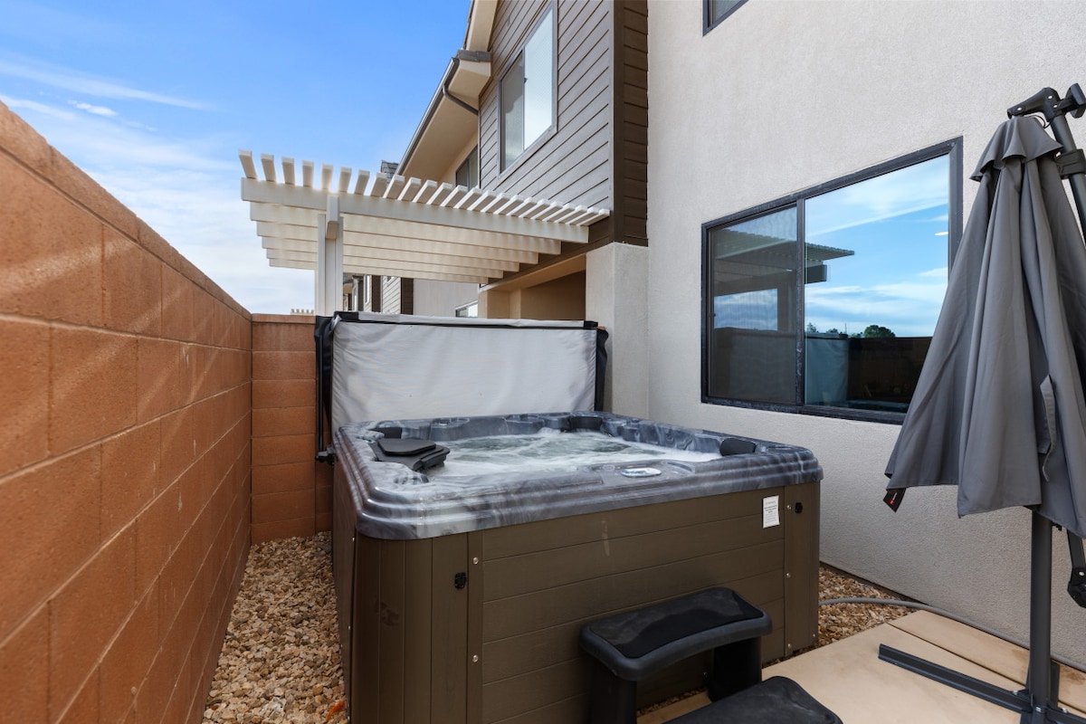 A private hot tub is situated in a secluded outdoor space, surrounded by a low wall for added privacy. A partially covered patio area provides shade, and a couple of black steps lead up to the tub, inviting relaxation in this tranquil setting.