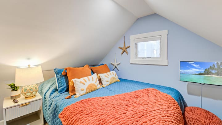 Acércate a este encantador dormitorio en el ático 2, completo con una cómoda cama, ropa de cama vibrante y un televisor para un refugio perfecto después de un día de playa.