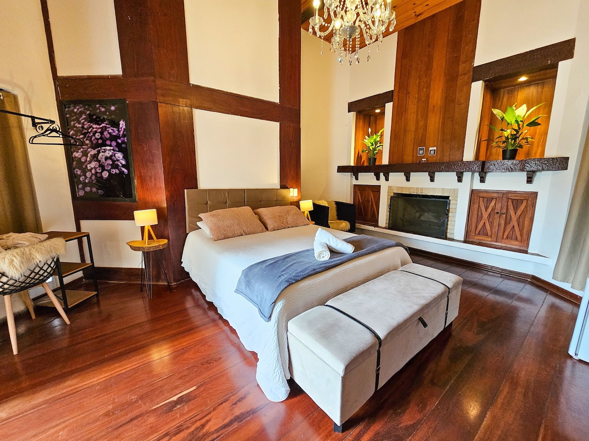 The sleeping area features a spacious bed adorned with neutral tones. A decorative bench sits at the foot of the bed. Soft lighting is provided by two bedside lamps. A cozy armchair and a decorative fireplace are included, with plants enhancing the ambiance.