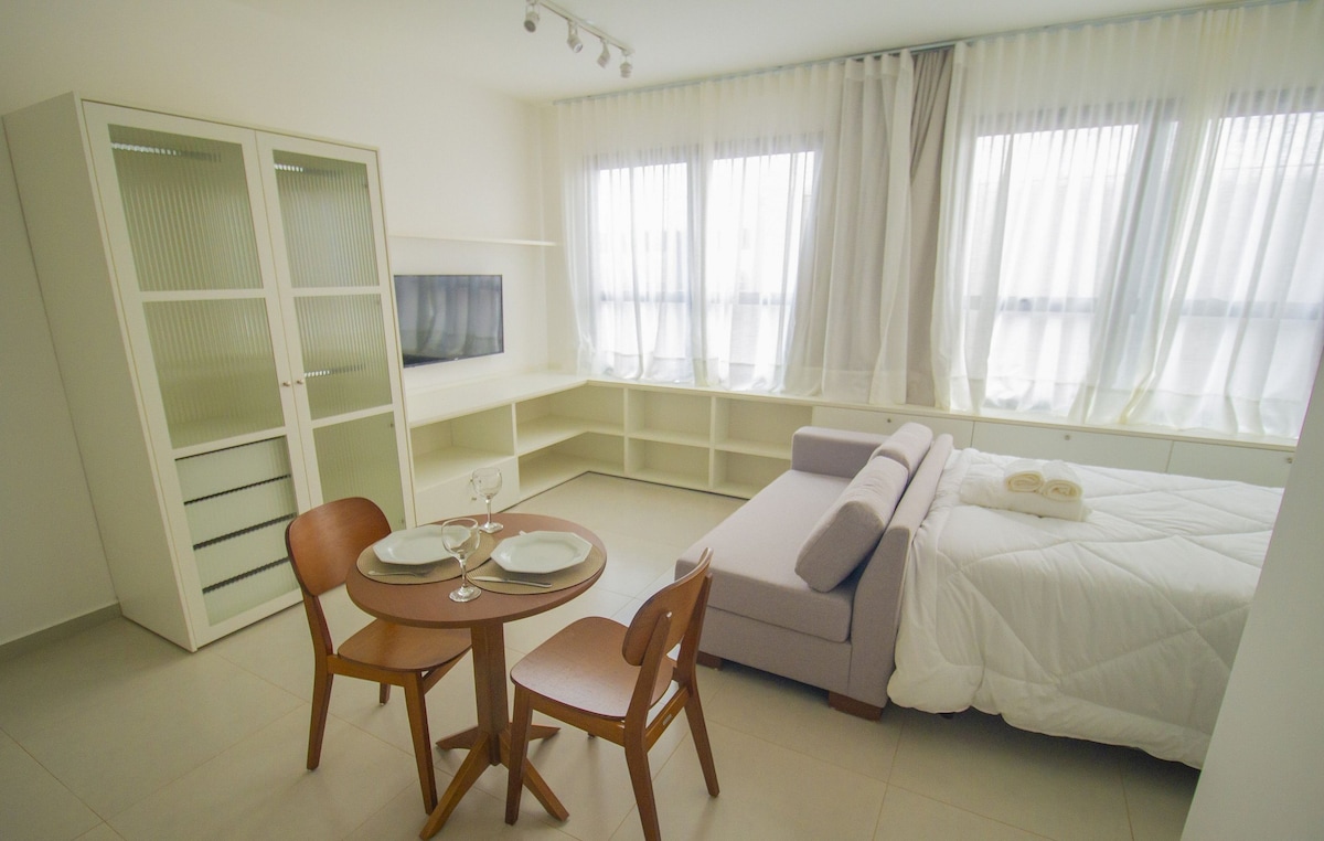 A light-filled living space features a compact kitchenette and dining area with a round table and two wooden chairs. A comfortable couch is positioned near a wall-mounted television. Large windows dressed in sheer curtains provide abundant natural light, enhancing the minimalist decor.