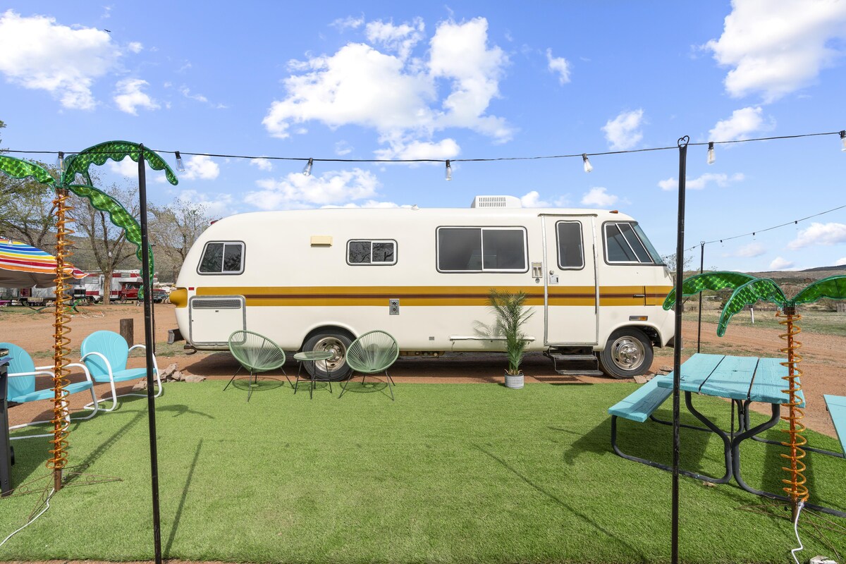 A charming vintage motorhome is positioned under a clear blue sky, surrounded by decorative palm trees. Outdoor seating includes two green lounge chairs and a picnic table set upon a grassy area, illuminated by string lights above, offering a welcoming space for relaxation.