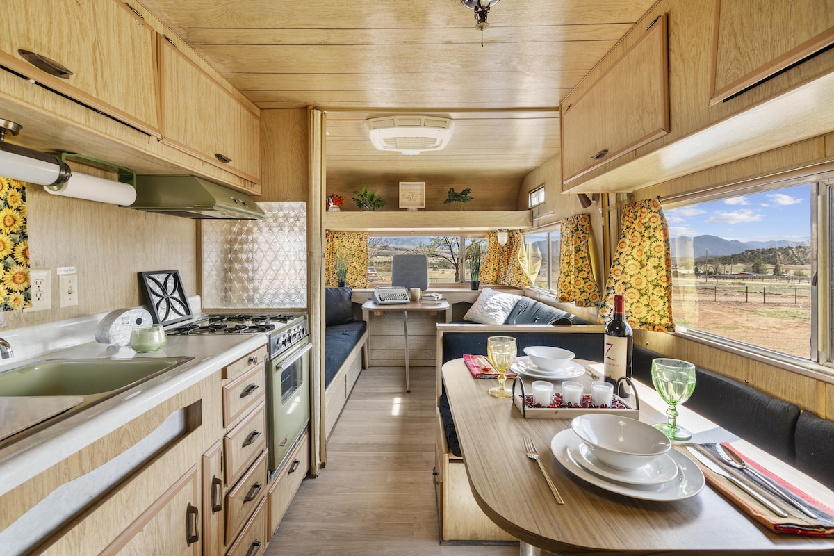 The interior of a vintage camper is displayed, featuring a kitchen area with a stove and sink alongside a dining table set for a meal. Large windows with floral curtains provide natural light and a view of the landscape, while cushioned seating offers comfort.
