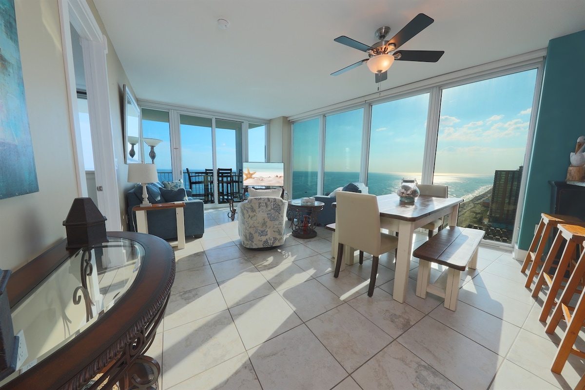 An open living space is illuminated with natural light, highlighted by floor-to-ceiling windows offering expansive views of the Gulf. A comfortable sofa and chair are positioned nearby, alongside a dining table that accommodates eight guests. A ceiling fan adds to the airy atmosphere.