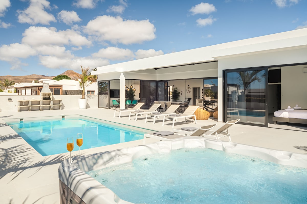 The outdoor area features a heated pool and a hot tub, both surrounded by sun loungers. An al-fresco dining area is visible, with refreshments placed on a nearby table. Modern architecture and expansive sliding glass doors create a seamless connection to the interior spaces.