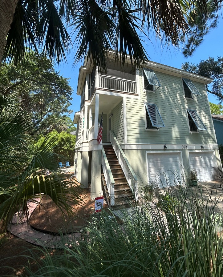 A two-story home is surrounded by lush greenery and tall palm trees. The light green exterior is complemented by white trim, and a U.S. flag is displayed near the entrance steps. The ground level features a garage, while a pathway leads to the front door.
