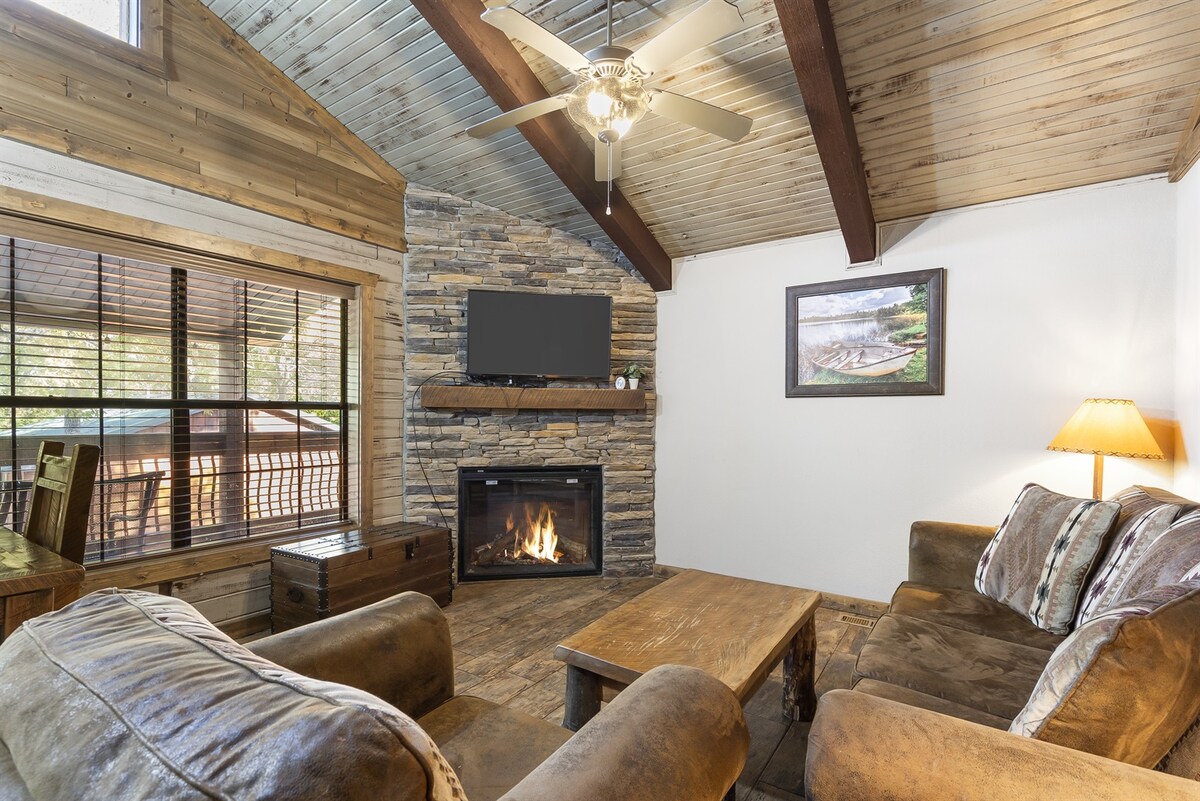 The living area presents a cozy setting with rustic wooden beams overhead and a stone fireplace as a focal point. Plush seating surrounds a natural wood coffee table. A flat-screen TV is mounted above the fireplace, and natural light filters through the large window.