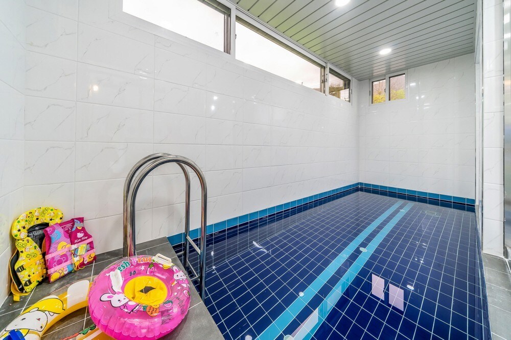 An indoor private pool is presented, featuring a tiled floor in shades of blue. The pool is equipped with a stainless steel ladder for easy access. Colorful inflatable toys are arranged on the poolside, providing a playful atmosphere.