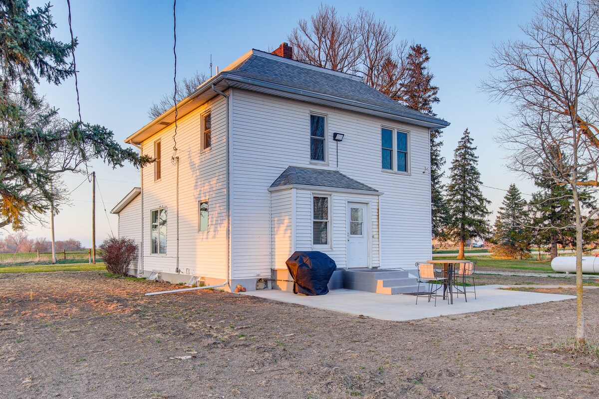 The exterior of a two-story house is shown, featuring a concrete patio with outdoor furniture and a gas grill. The property is surrounded by grassy areas and a few trees, with distant fields visible. Soft evening light adds warmth to the welcoming exterior.