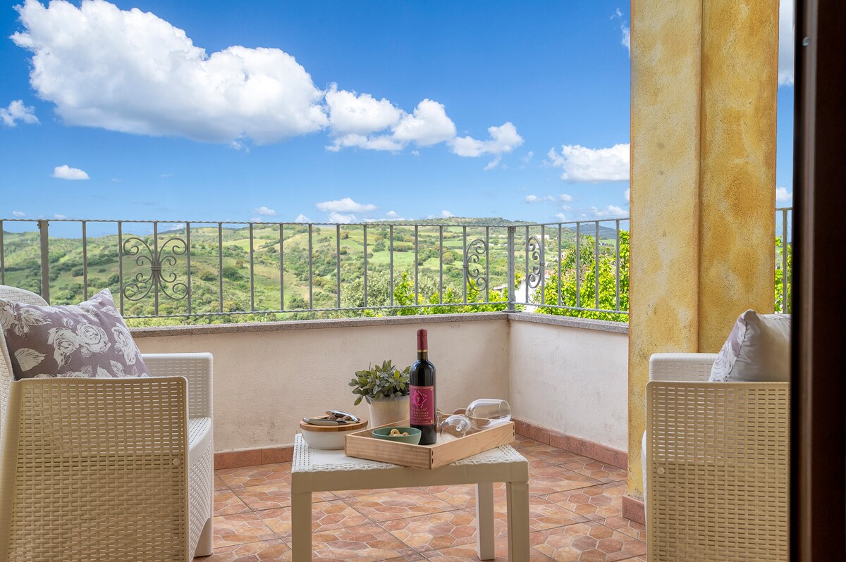 A private balcony is presented with two comfortable chairs facing a scenic view of green hills under a blue sky. A small table holds a bottle of wine and a tray with snacks, inviting relaxation in a serene outdoor setting.