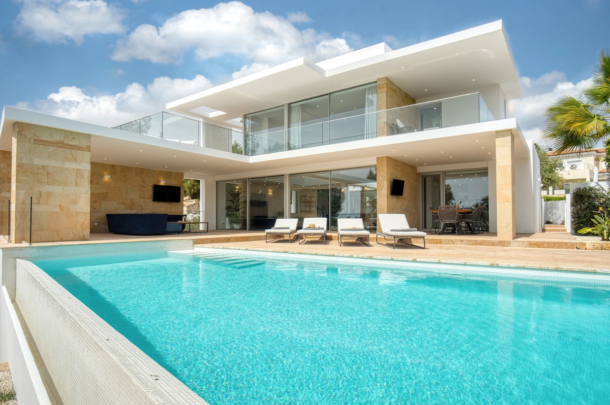 The villa features a modern two-story design with large glass panels that provide views of the pool area. Loungers are placed beside the inviting blue pool, surrounded by a landscaped outdoor space. A spacious terrace offers additional relaxation areas, enhancing the overall setting.