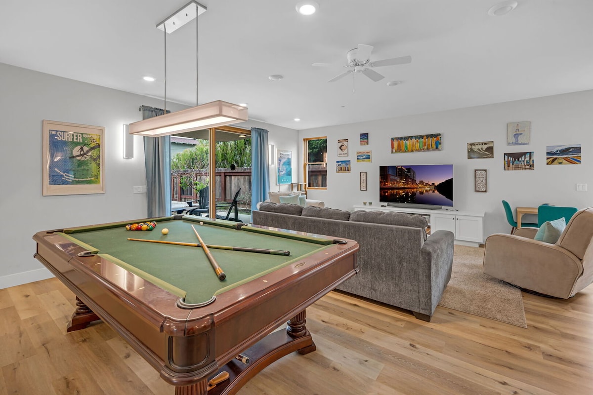 An entertainment area features a pool table with cues resting on the side. A smart TV is mounted on the wall, displaying various images. Comfortable seating is arranged, and large sliding doors provide access to an outdoor space with greenery.