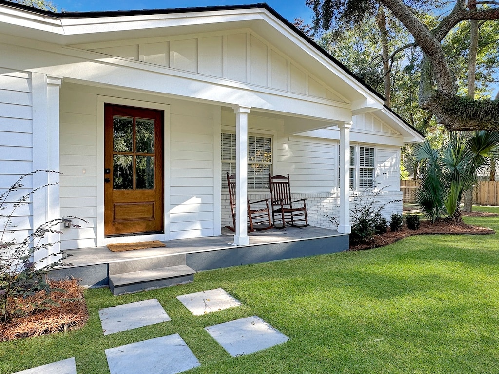 The exterior of a charming one-story house is illuminated by natural light. A welcoming entryway features a classic wooden door and two rocking chairs on the porch, while neatly trimmed grass and stone walkways lead to the entrance. Lush greenery surrounds the property.