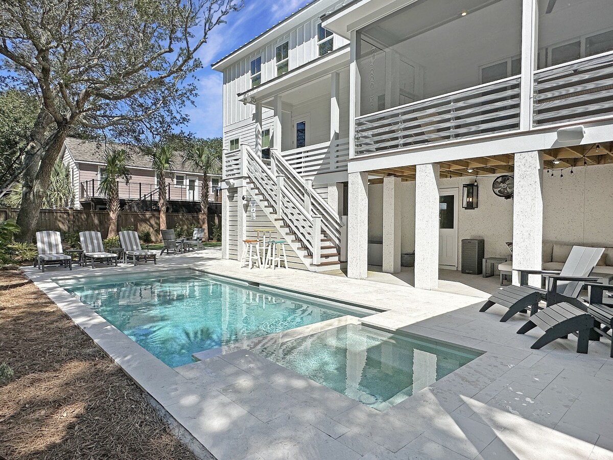 An outdoor space features a private pool with a shallow area and sun loungers arranged around it. A covered area is visible, providing shade, while stairs lead up to the elevated house. Lush greenery surrounds the backyard, enhancing the serene environment.