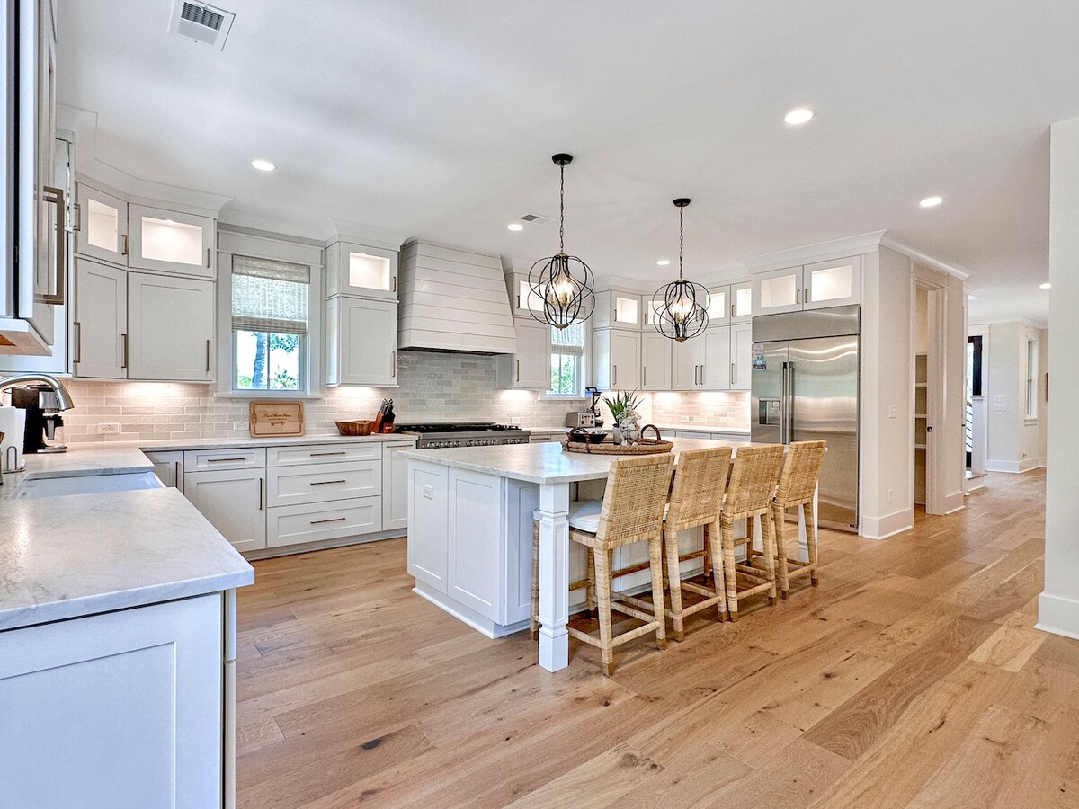 A spacious kitchen features white cabinetry and a large central island with seating for four. Pendant lights accentuate the area above the island. Stainless steel appliances and a tiled backsplash enhance the modern design, while natural light brightens the room through multiple windows.