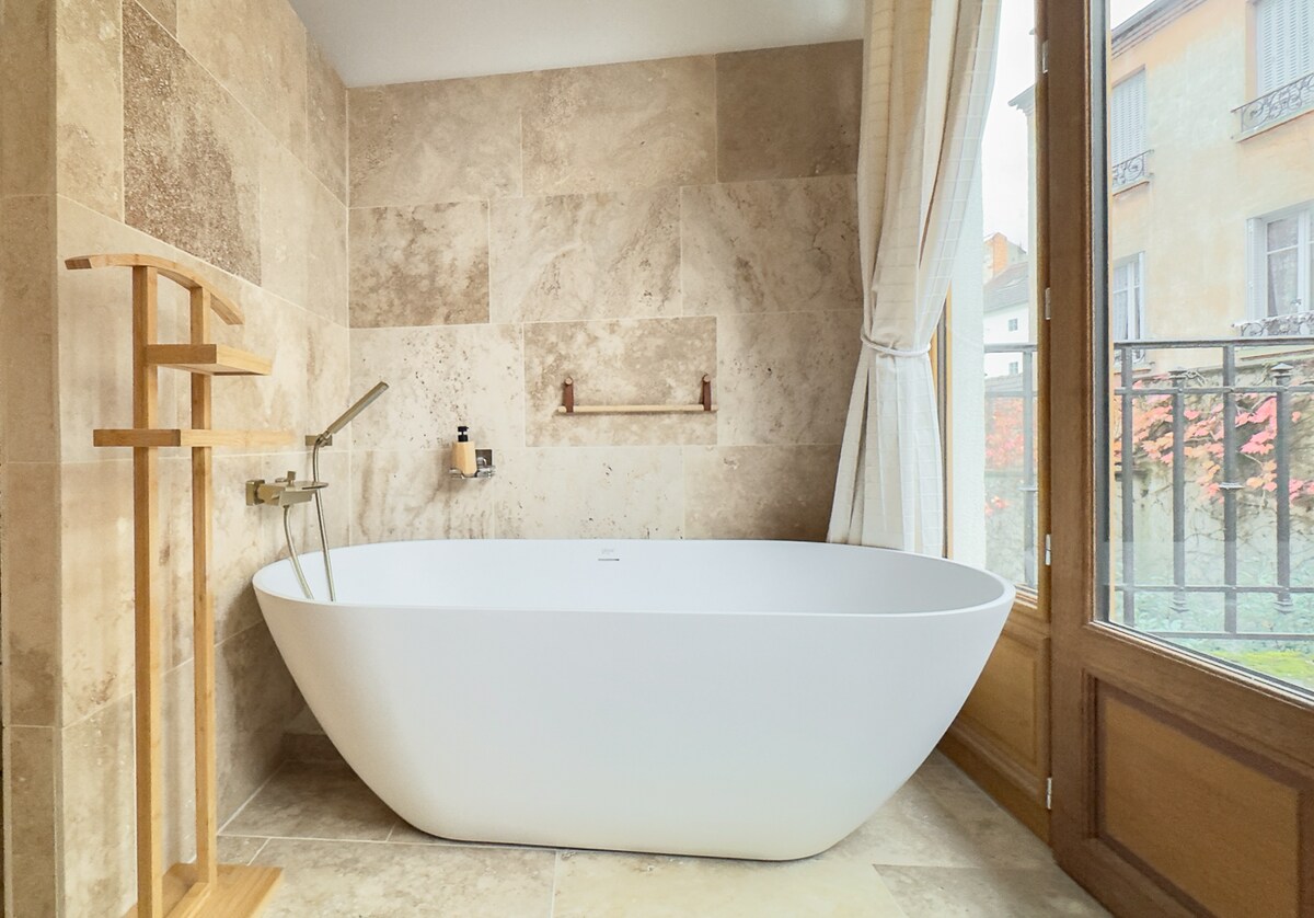 A modern freestanding bathtub is positioned against a wall of natural stone tiles, creating a soothing atmosphere. Soft light filters through a window, with a wooden rack beside the tub, providing space for toiletries. The space is accented by elegant window treatments.
