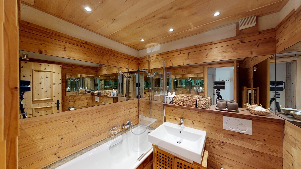A spacious bathroom features wooden paneling and a combination of warm lighting. A bathtub with a shower is positioned beside a modern sink set within a wooden cabinet. Mirrored surfaces enhance the sense of space, while neatly arranged toiletries add convenience.