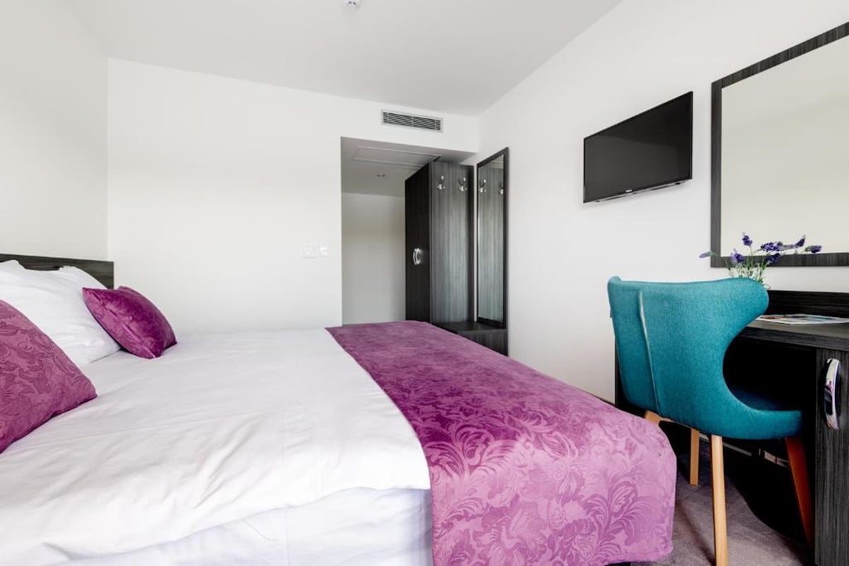 A modern bedroom features a large bed dressed in crisp white linens with a decorative purple throw. A chair in a vibrant teal color is positioned at a sleek desk. A flat-screen TV is mounted on the wall, and double doors lead to an adjoining space.