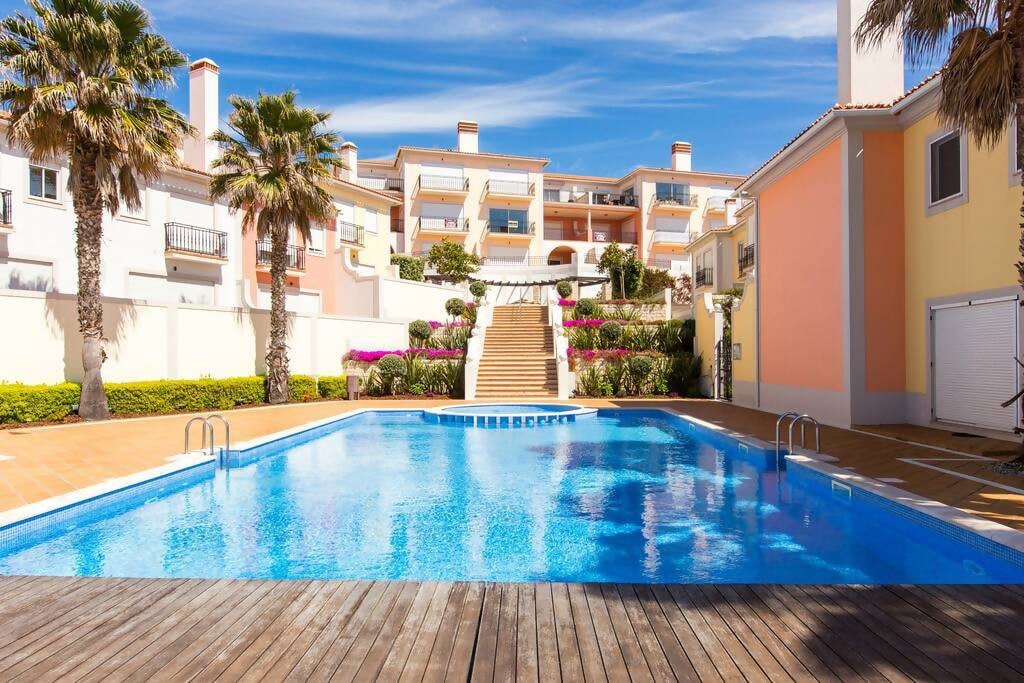 A spacious outdoor swimming pool is framed by palm trees and colorful flowering plants. Surrounding the pool area are well-maintained residential buildings, with stairs leading up to a landscaped terrace. The vibrant blue water reflects the clear sky above, inviting relaxation.