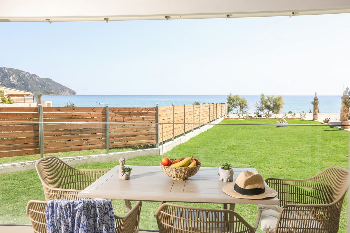 A dining table is set for outdoor meals, surrounded by comfortable chairs. A bowl of colorful fruit sits at the center. The expansive view includes a green lawn leading to the sea, framed by wooden fencing and distant hills.