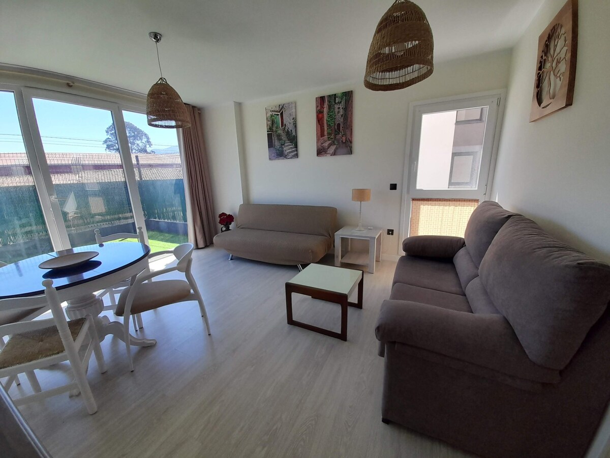 The living room features two comfortable sofas, one of which is a sofa bed. A circular dining table with chairs stands beside the large windows, allowing natural light to fill the space. Artistic wall decor adds character, while a door leads to a terrace.