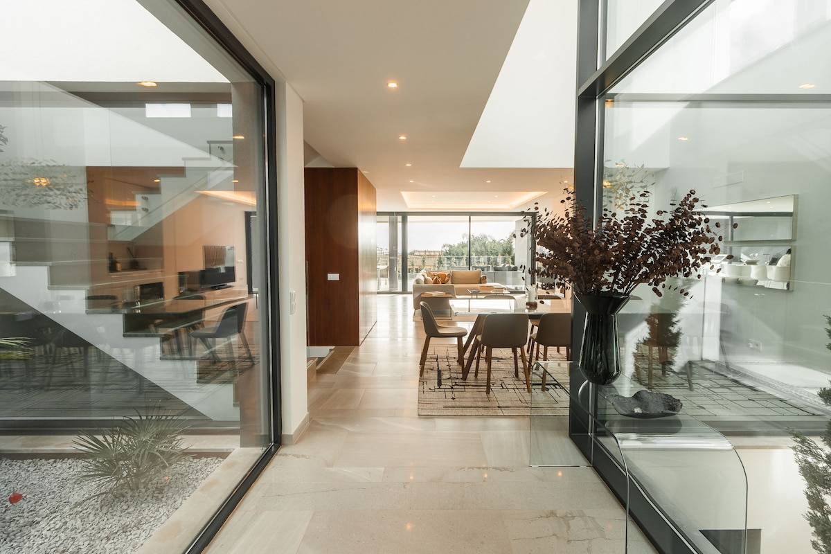 An open entrance showcases floor-to-ceiling glass walls, allowing natural light to fill the space. The modern interior features a blend of dining and living areas, with stylish furniture and decor. A lush garden is visible through the glass, enhancing the tranquil ambiance.