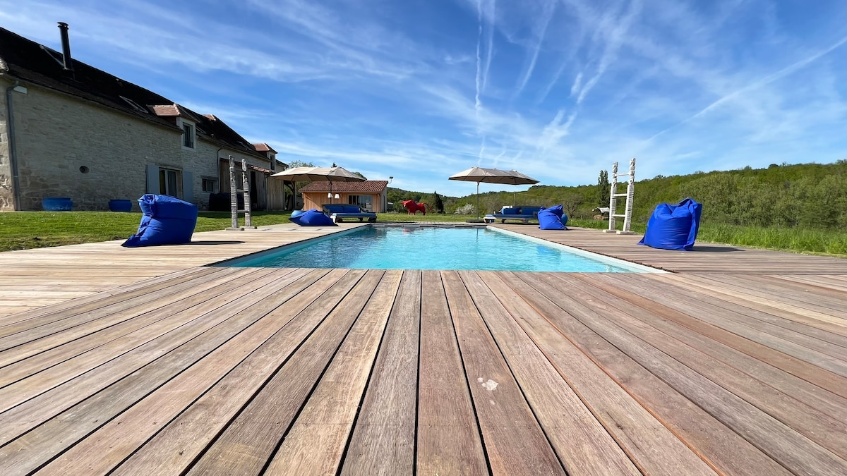 A spacious wooden deck surrounds a private swimming pool, featuring a clear blue water surface. Bright blue bean bag chairs are placed on the deck, while umbrella shades provide relief from the sun. Lush green fields are visible in the background under a clear sky.