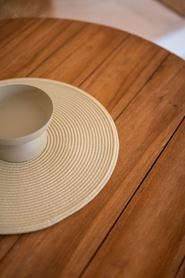 A wooden table surface features a round woven placemat in a neutral tone, with a simple, light-colored bowl positioned in the center. The warm wood grain is visible, creating a natural ambiance.