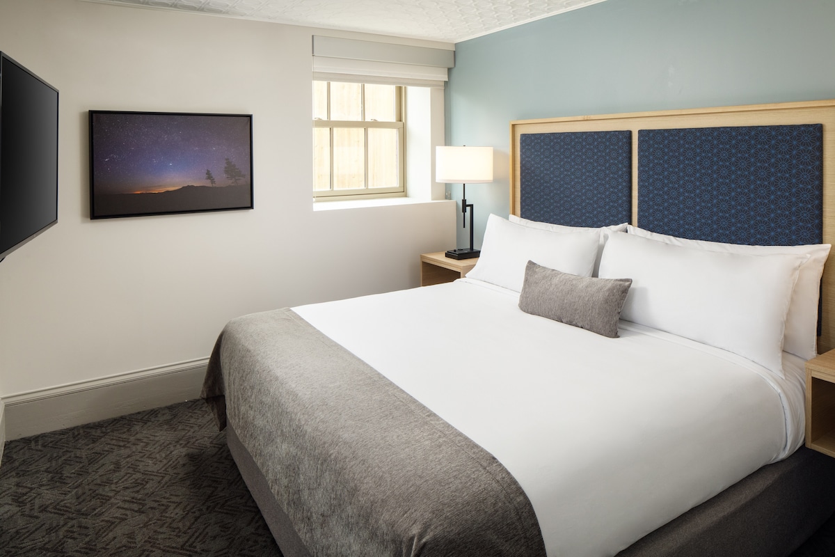 A spacious guest room features a king bed with a light-colored duvet and a decorative pillow. A bedside lamp stands on a nightstand, while a 55-inch TV is mounted on the wall opposite. A large window allows natural light to fill the space.