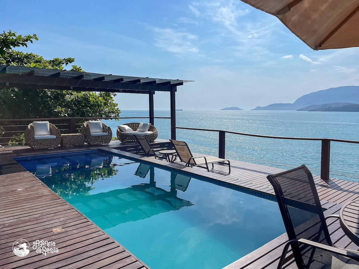 A serene outdoor area features a swimming pool surrounded by wooden decking, with sun loungers arranged for relaxation. Lush greenery provides shade, and the landscape includes a stunning view of the ocean, with distant hills visible under a partly cloudy sky.