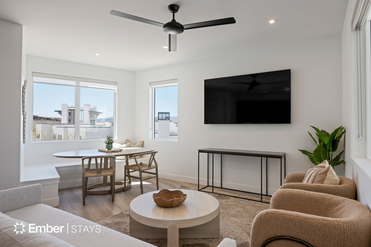 A modern living area features a comfortable seating arrangement, including plush armchairs and a round coffee table. Large windows allow natural light to fill the space, while a dining table is positioned nearby. A flat-screen TV is mounted on the wall opposite the seating.