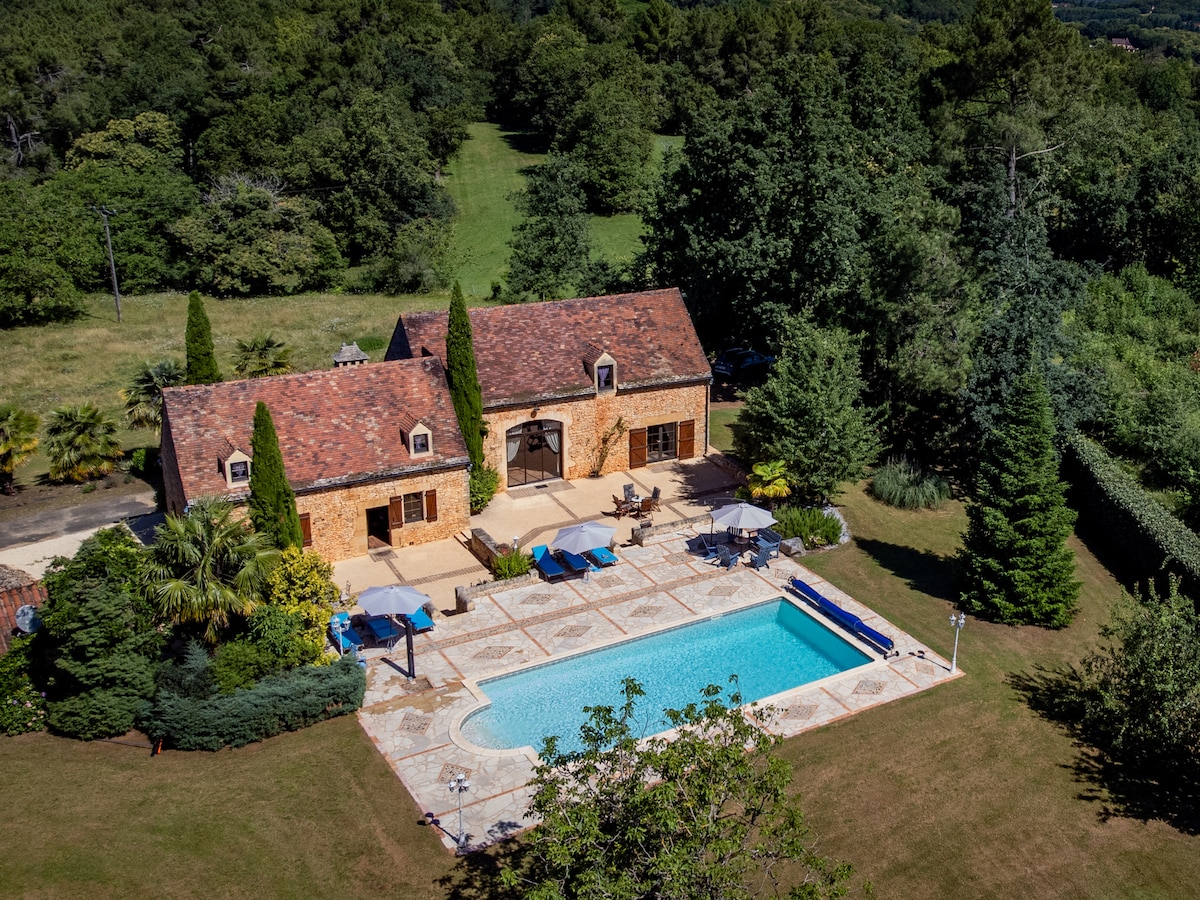 An aerial view of a charming 17th-century house surrounded by lush greenery. A private swimming pool is situated in front, complemented by sun loungers and umbrellas on the terrace. The building features a rustic stone exterior and a traditional tiled roof, surrounded by well-maintained gardens.