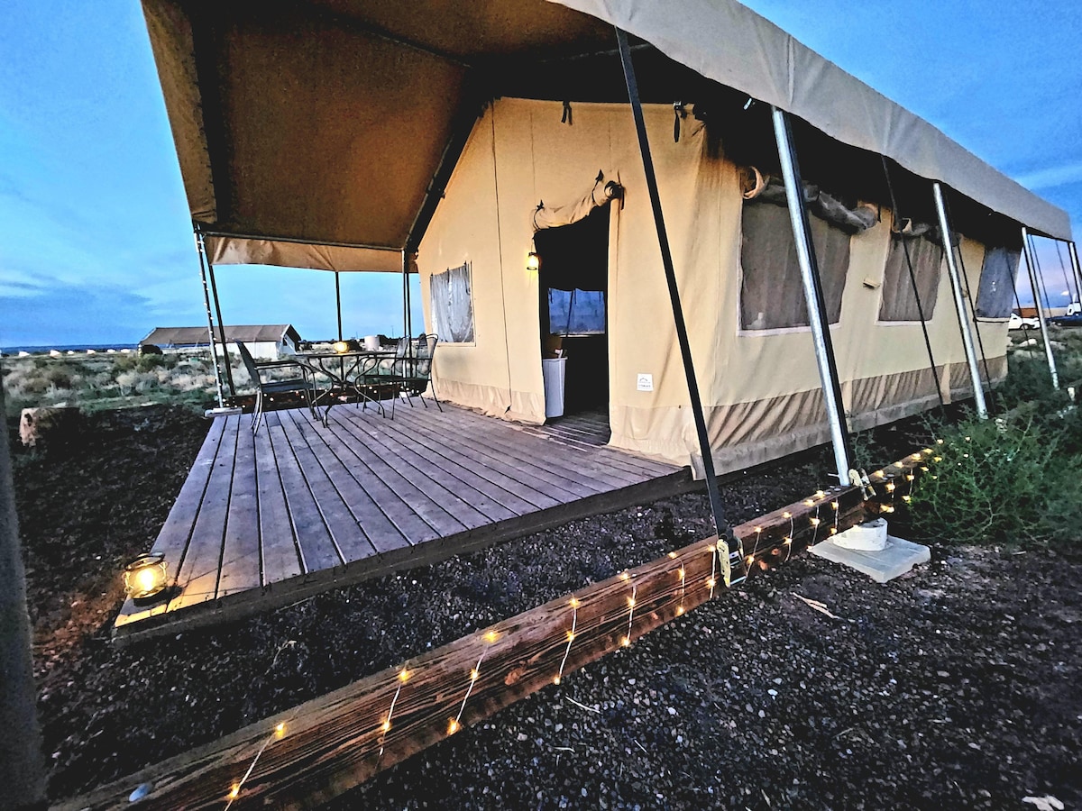 The family tent features a spacious wooden deck with soft outdoor lighting. A table and chairs are provided outside, ideal for enjoying the surrounding landscape. The tent's sturdy construction is complemented by large windows for natural light and ventilation.