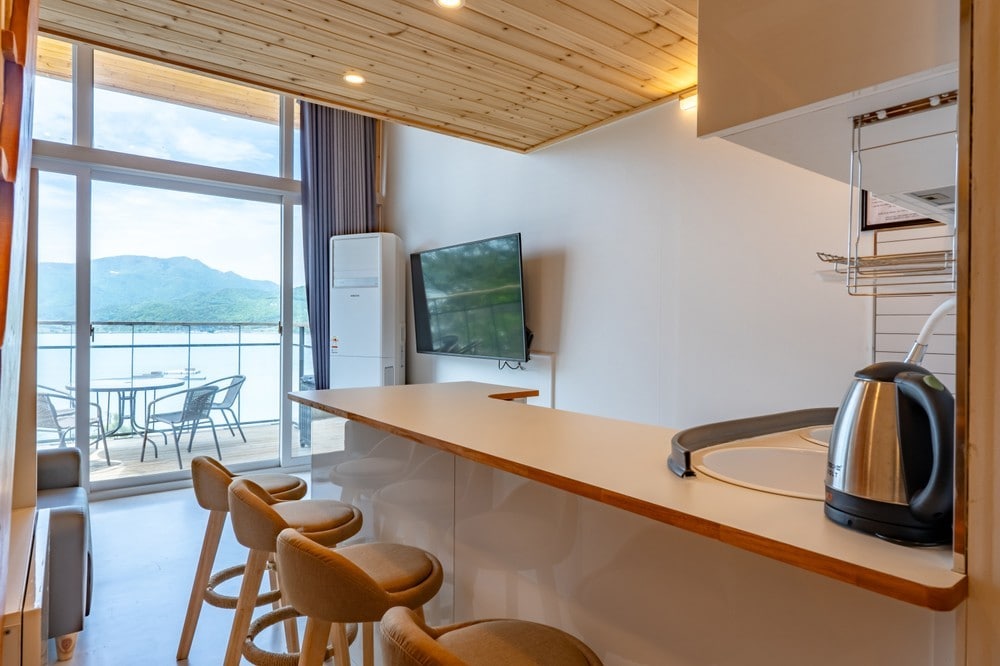 An open kitchen area features a sleek countertop with wooden stools, offering a view of the outdoor seating and scenic mountains. A flat-screen television is mounted on the wall, while a kettle sits on the countertop, adding functionality to the space.