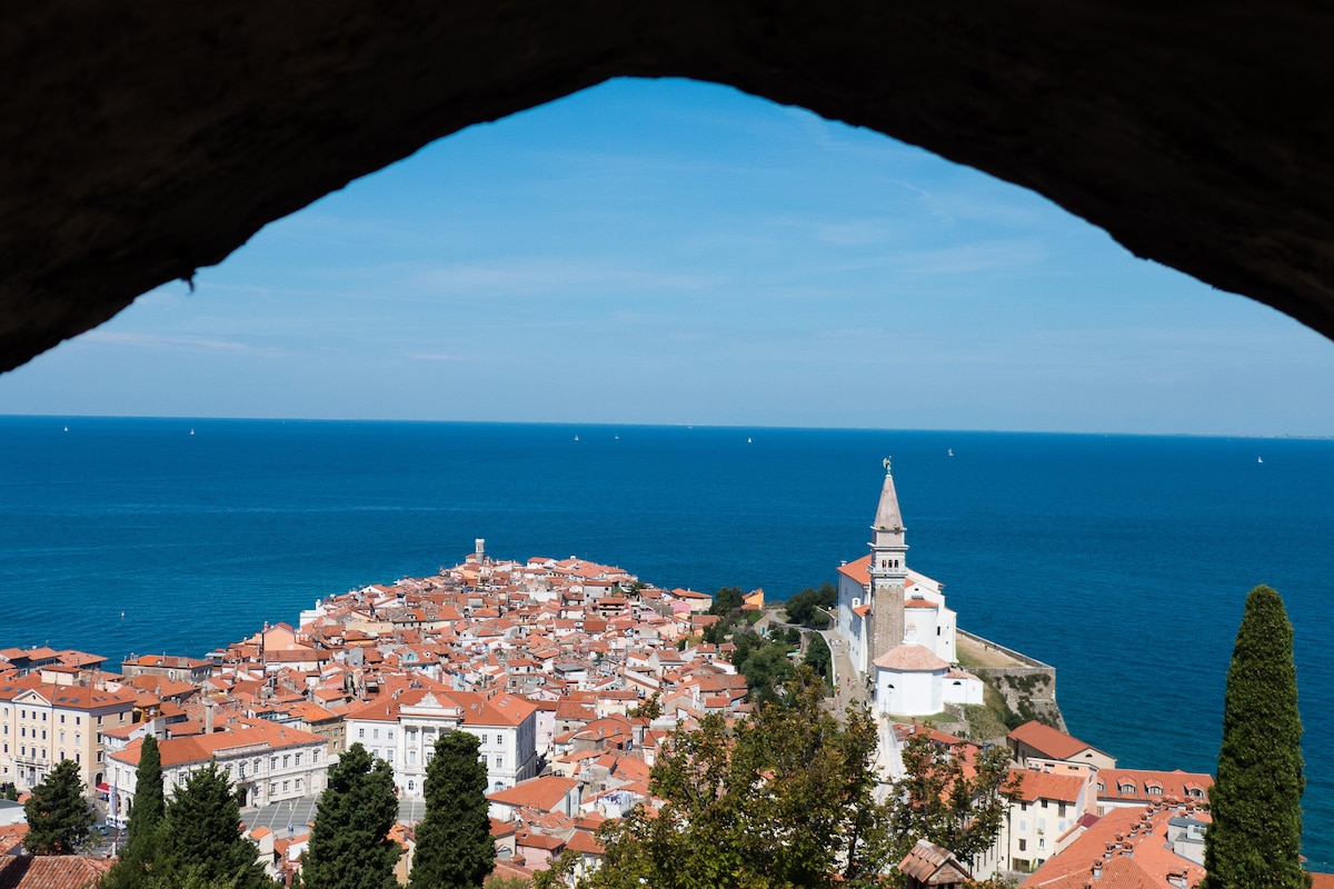 A panoramic view of a coastal town is framed by an archway. The vibrant rooftops and historic buildings are seen nestled against the azure waters of the sea, while a prominent church tower adds to the picturesque scene.
