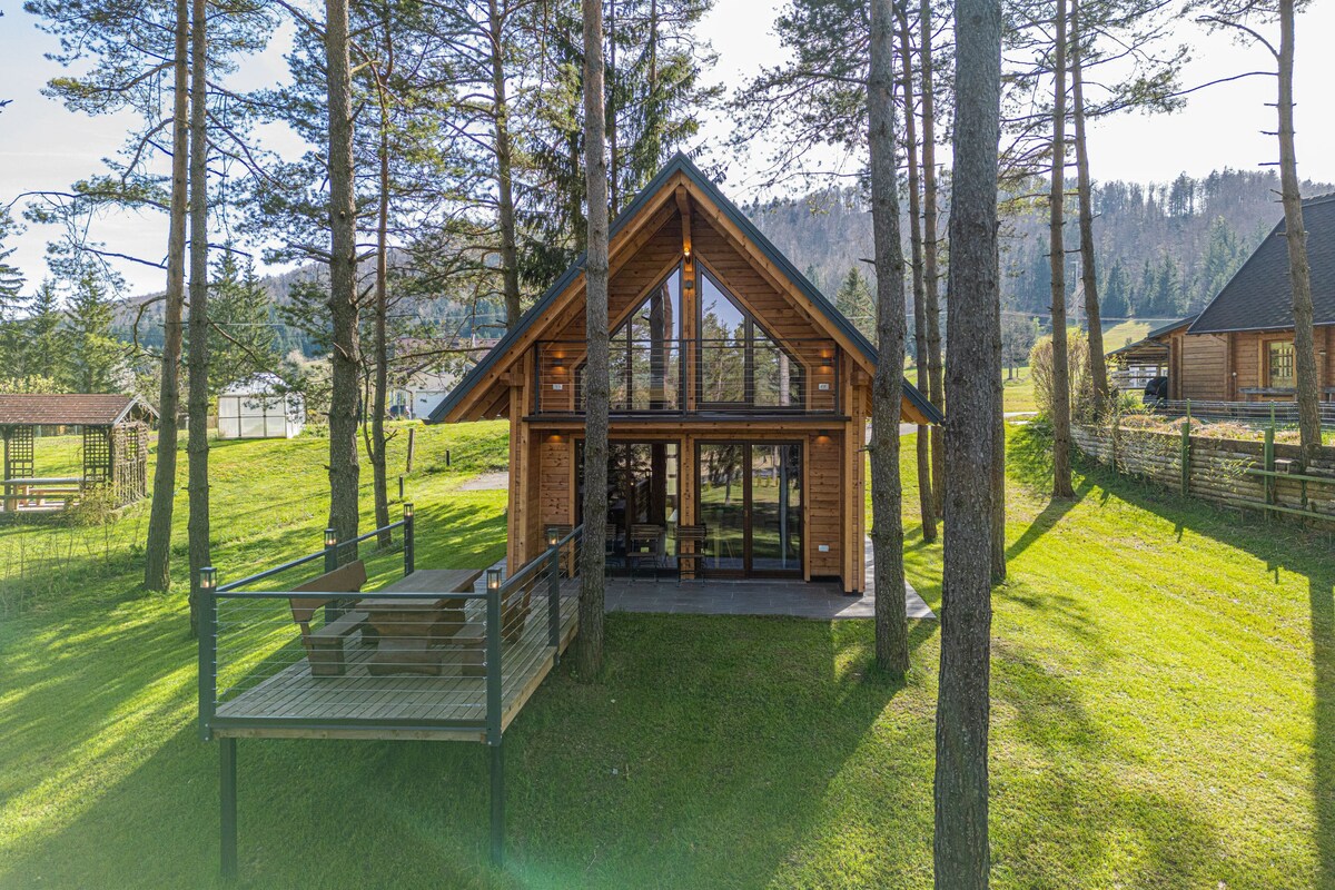 The contemporary chalet is surrounded by tall trees and open grassy areas. A private deck with a picnic table and benches extends from the house, inviting outdoor relaxation. Large windows reveal the warm wood paneling of the interior, blending harmoniously with the natural surroundings.