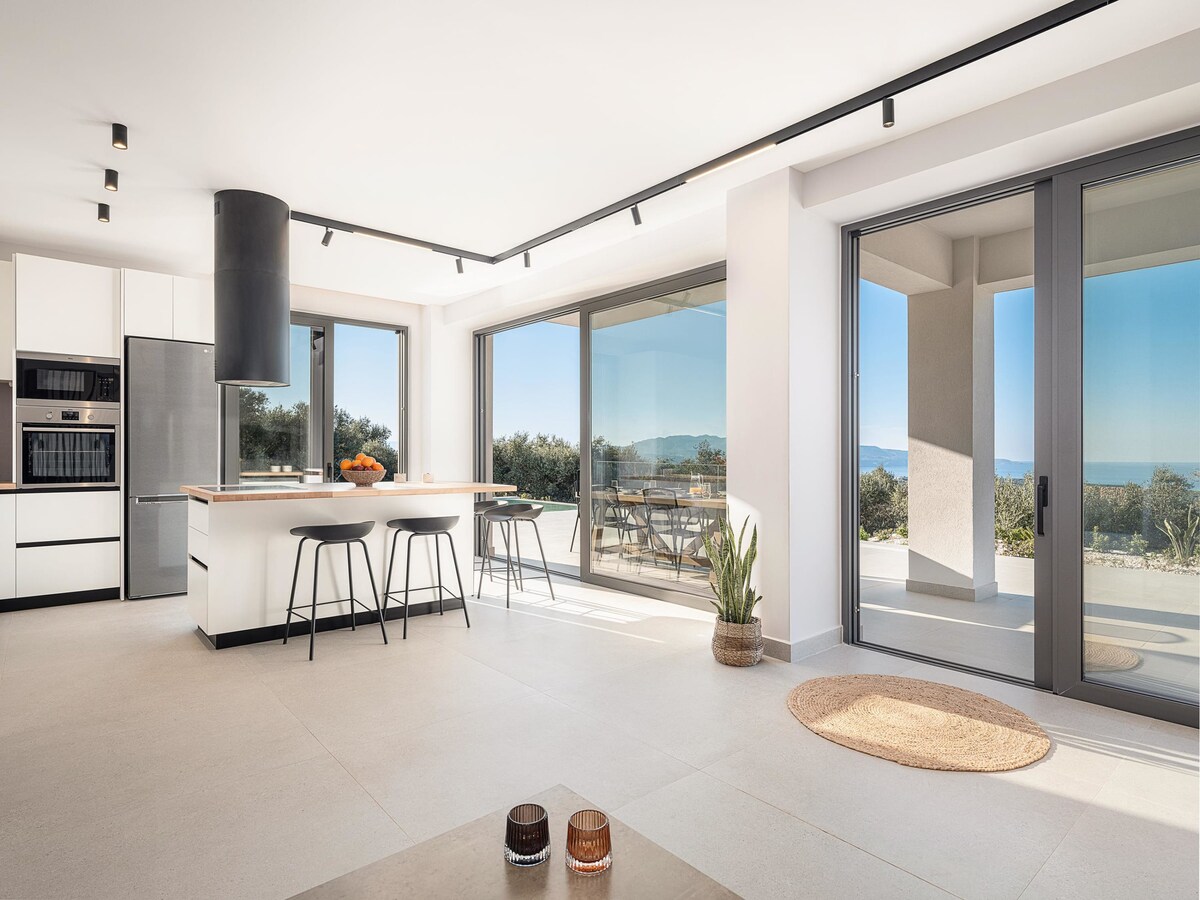 The modern kitchen features sleek cabinetry and an island with seating for three. Large glass doors reveal a bright outdoor terrace, enhancing the space with natural light and scenic views of the landscape. A light-colored rug is positioned on the tiled floor.