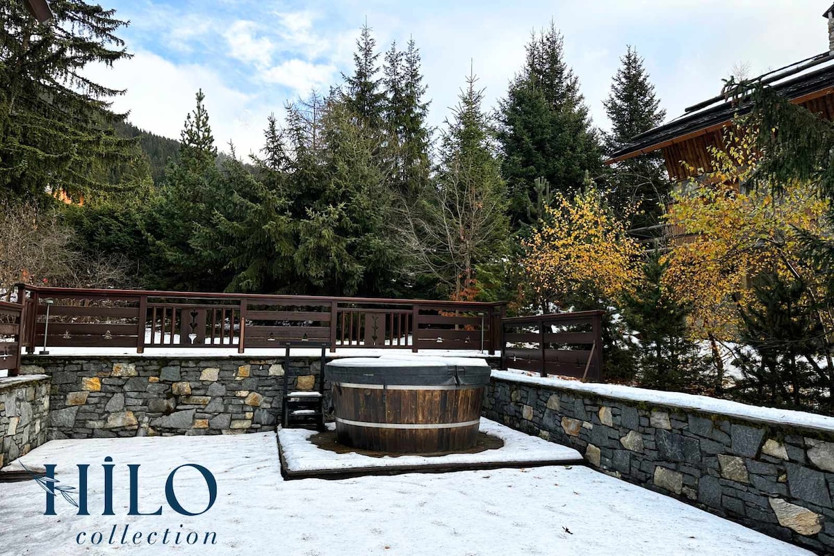 A wooden hot tub sits on a stone patio surrounded by a snow-covered landscape. Tall evergreen trees provide a natural backdrop, and a wooden railing outlines the area. The peaceful scene reflects a serene retreat in the heart of nature.