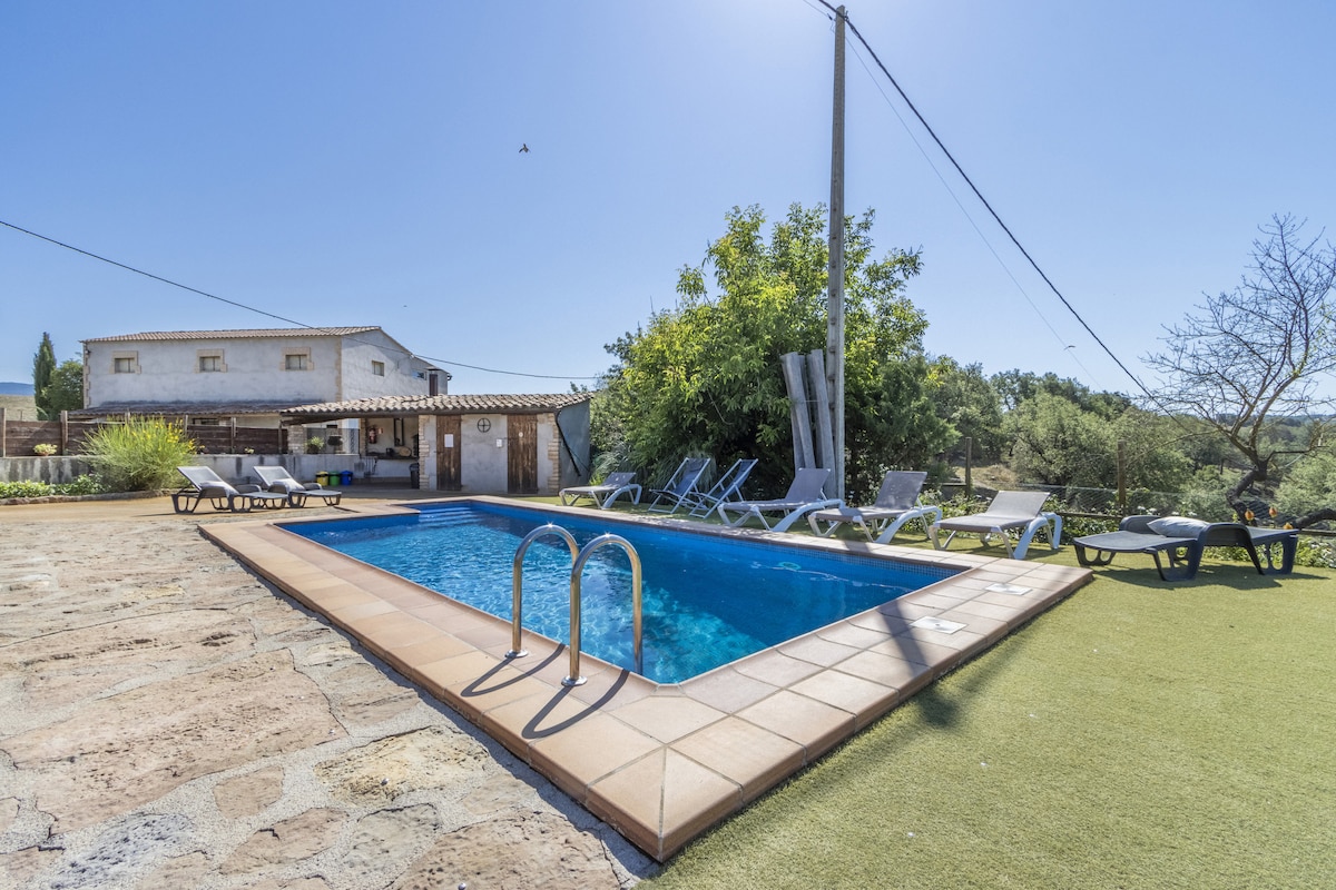A private outdoor space features a swimming pool surrounded by sun loungers. Greenery is visible nearby, and a rustic building is in the background. The area is illuminated by natural light from a clear blue sky.