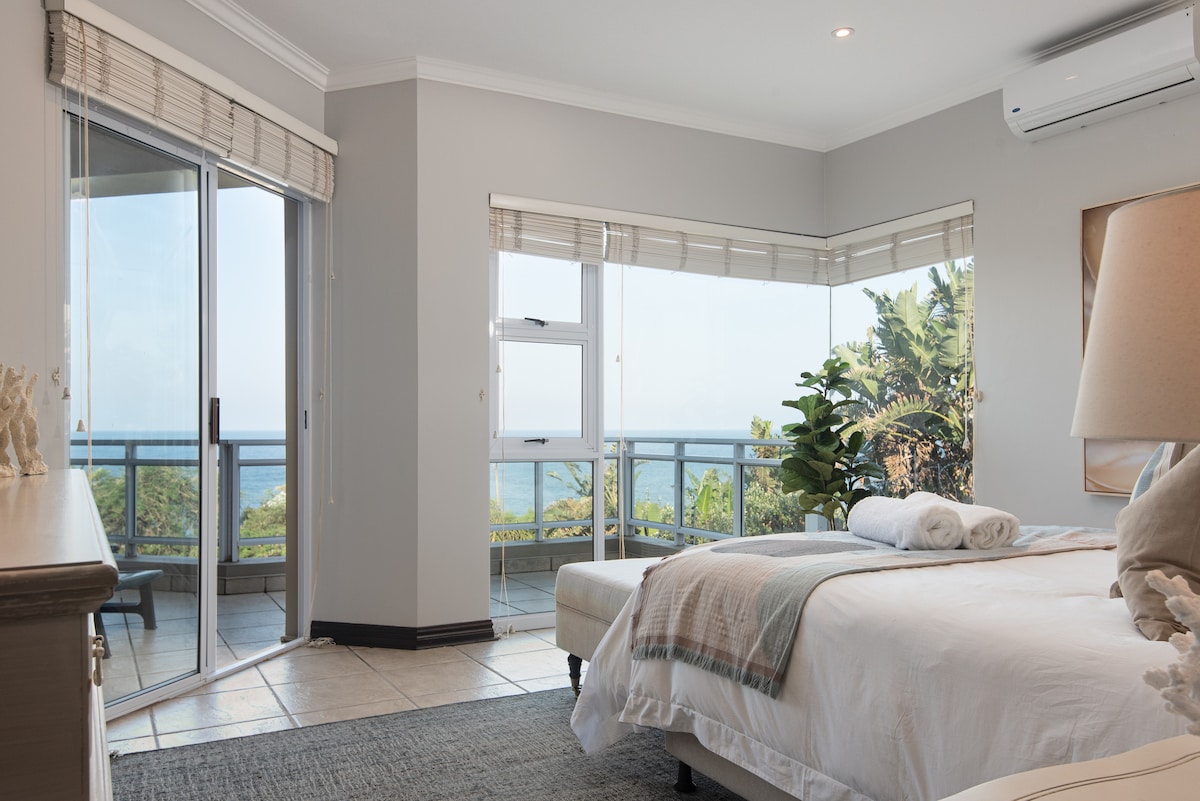 A well-lit bedroom features a spacious bed with neatly arranged pillows and a soft throw. Large glass doors open to a balcony, offering views of the ocean and surrounding greenery. Natural light enters through the windows, enhancing the fresh and modern decor.