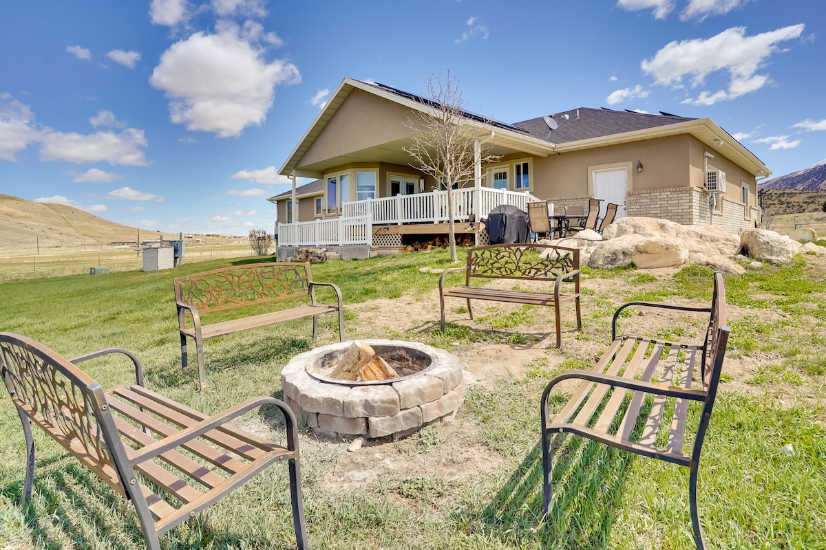 An outdoor seating area surrounds a stone fire pit, complemented by multiple metal benches. The beige-toned structure of the rental is visible in the background, featuring a spacious porch. Lush green grass and rolling hills under a blue sky complete the scenic landscape.