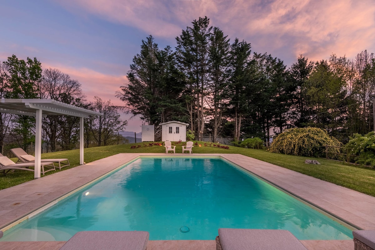A serene private pool framed by grassy lawns and trees stretches out beneath a vibrant sunset sky. Two lounge chairs edge the pool, while a covered seating area and a small cottage offer additional relaxation options amidst the tranquil landscape.