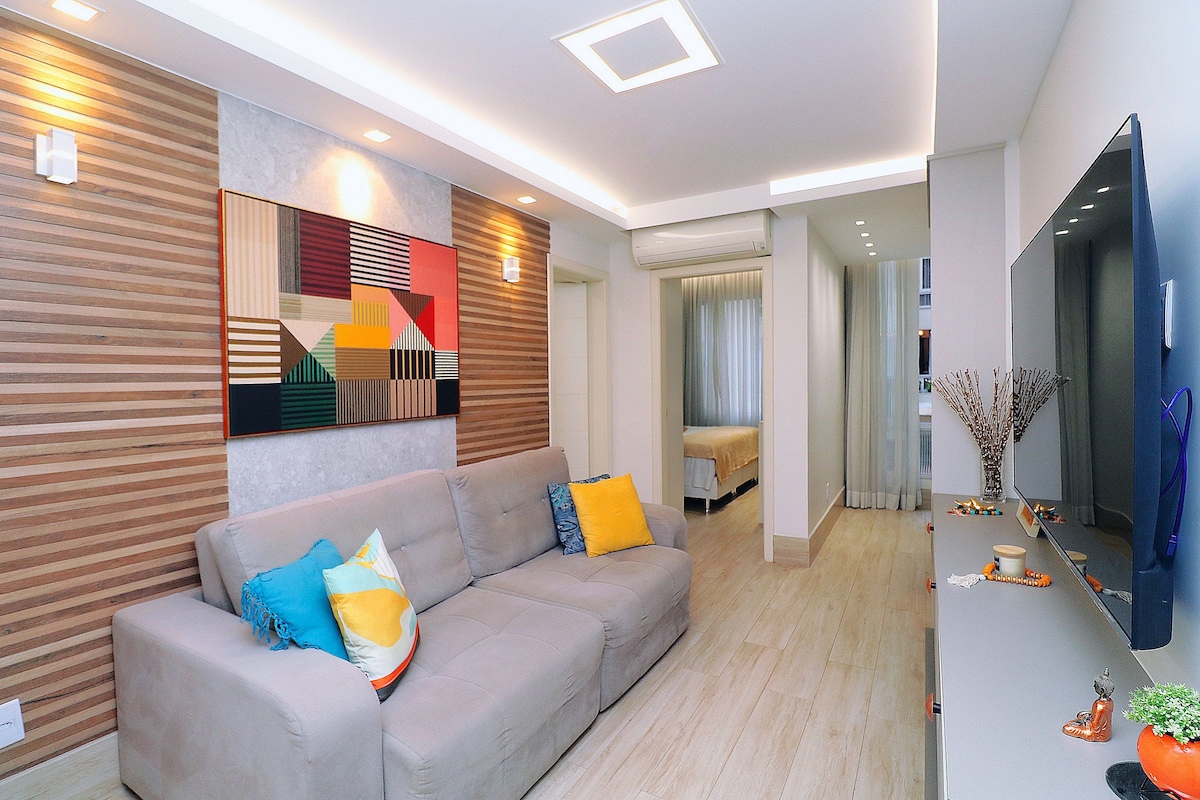 A modern living area features a comfortable gray sectional sofa adorned with colorful cushions. A large wall-mounted SmartTV is positioned opposite the seating, while artistic wall décor with geometric designs adds visual interest. Soft lighting enhances the inviting ambiance.