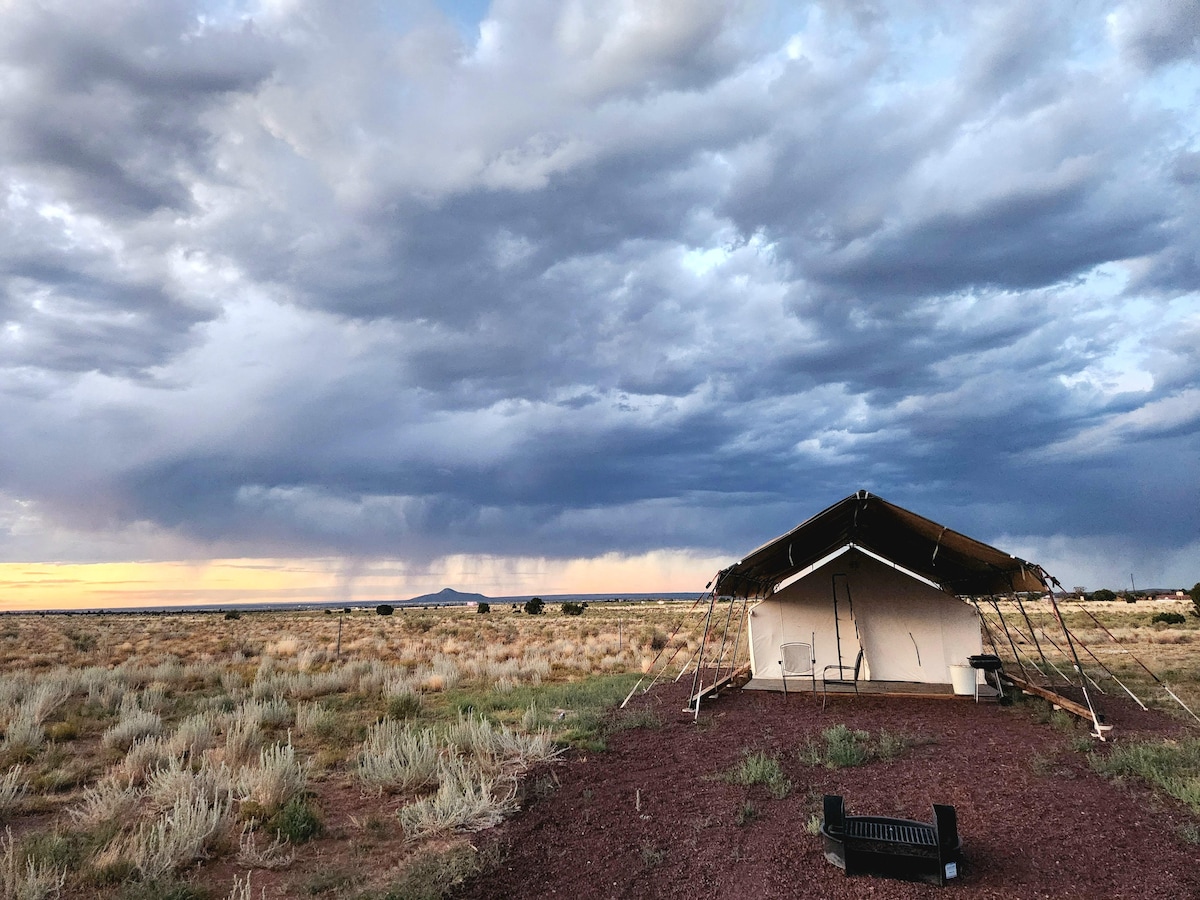 A cozy glamping tent is set against a vast prairie landscape. Tall grasses and scattered brush surround it, while dramatic clouds fill the sky, transitioning from gray to soft orange hues at the horizon. A campfire area is visible nearby.