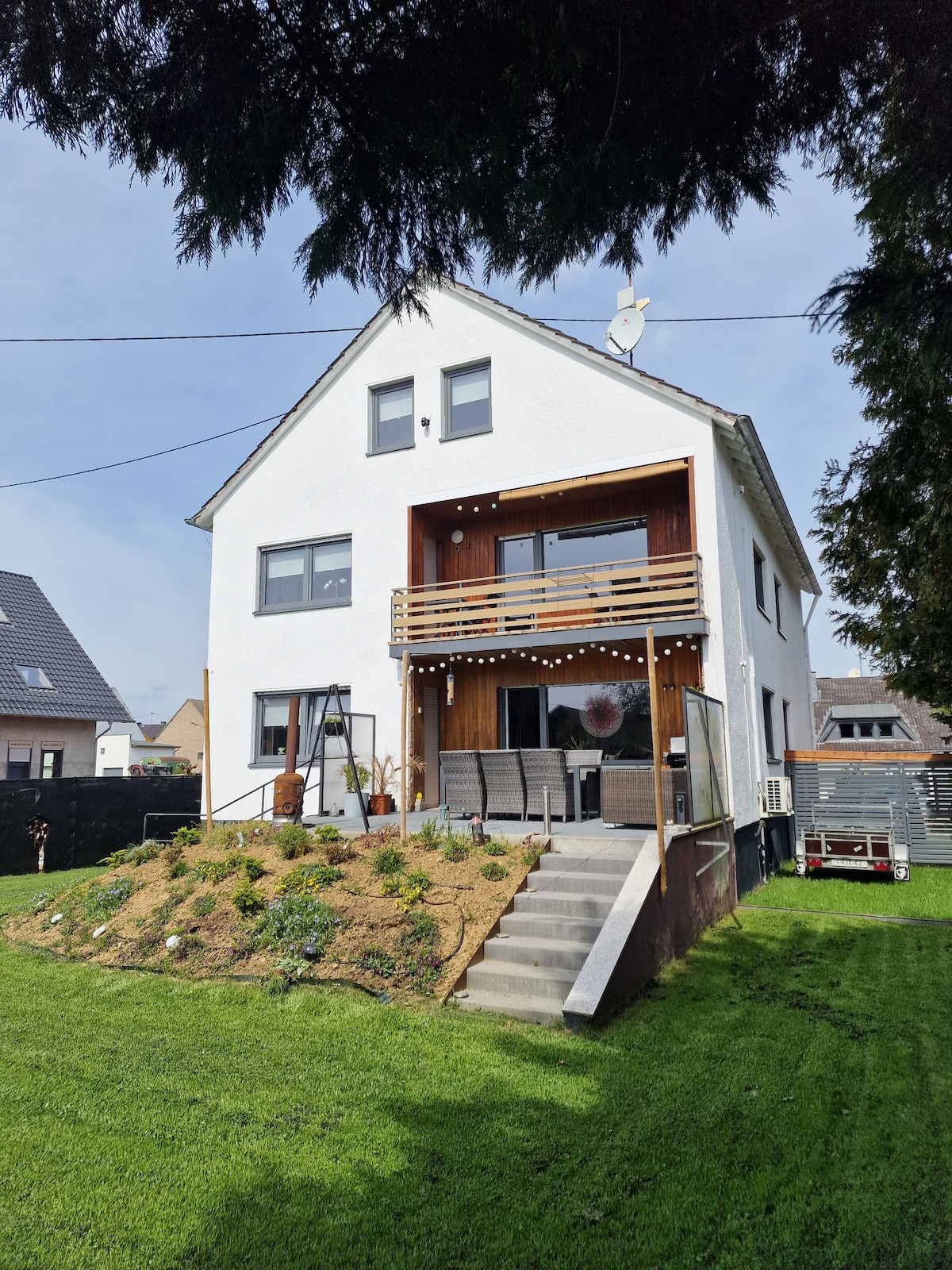 The exterior of the holiday apartment showcases a two-story house featuring a white façade. A wooden balcony is visible on the upper level, overlooking a landscaped garden. Steps lead down to a grassy area, with a seating arrangement on the terrace below.