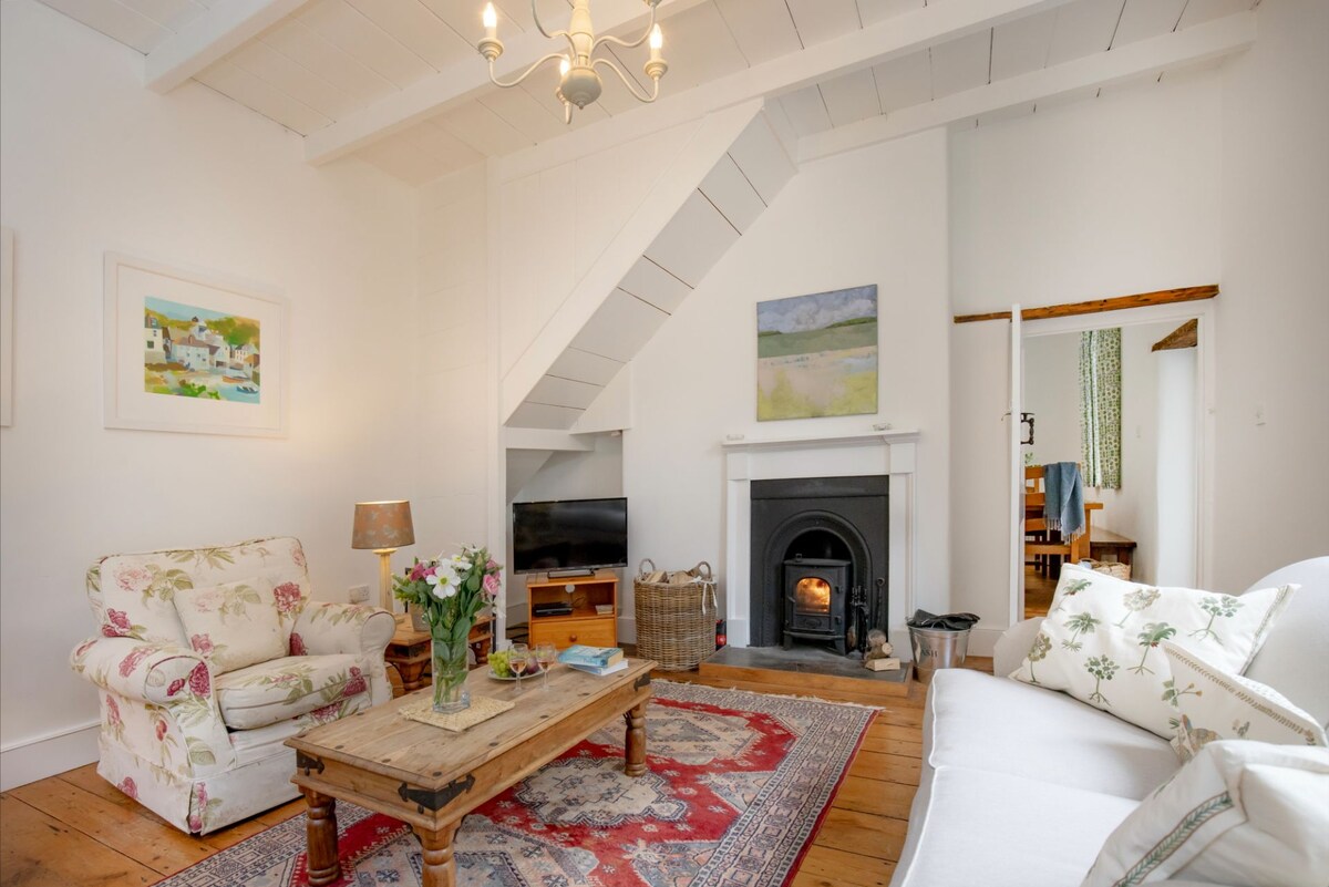 The living room features a wood-burning stove framed by a white mantel, with comfortable seating arranged around a rustic wooden coffee table. A floral armchair and light sofa complement the warm wooden floors, while art and fresh flowers enhance the inviting atmosphere.