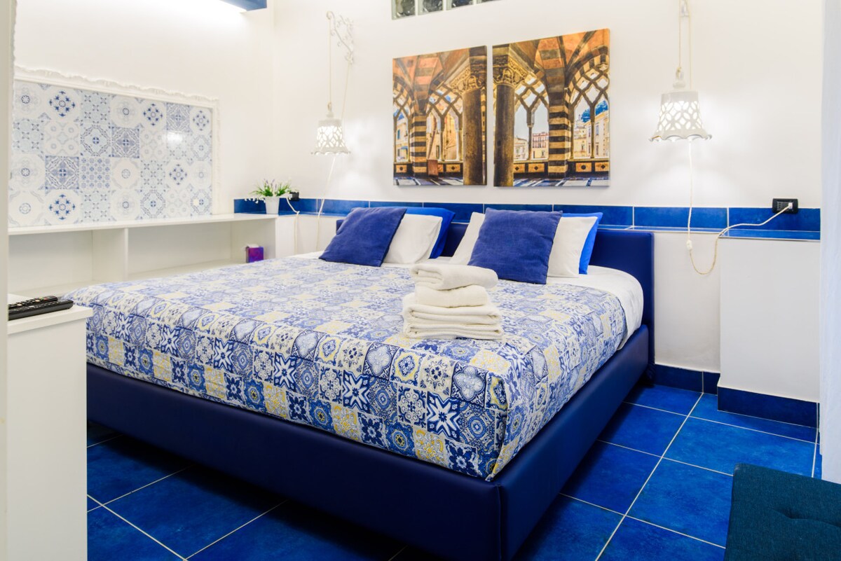 A spacious bed is adorned with a patterned blue and white coverlet, complemented by white and navy pillows. Two decorative wall lights hang symmetrically above the headboard. A piece of artwork featuring intricate architectural details is prominently displayed on the wall.