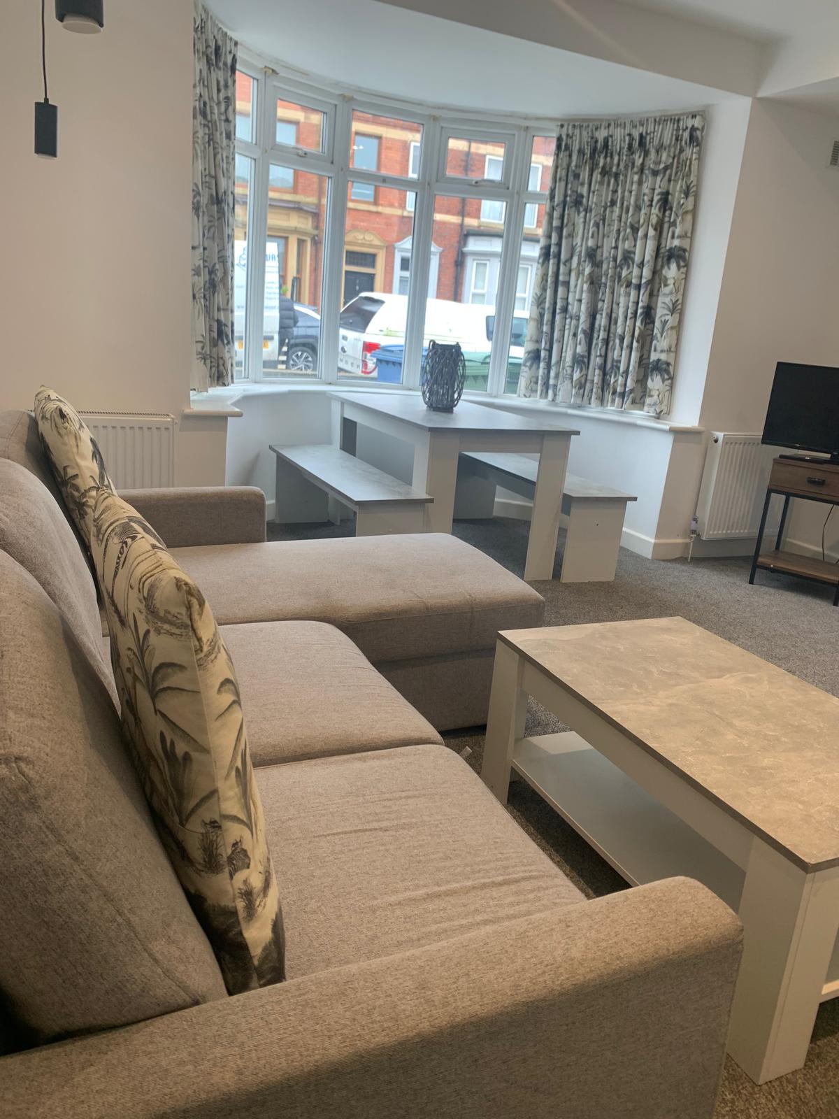 A comfortable seating area includes a gray sectional sofa with patterned cushions, facing a simple coffee table. Large windows allow natural light to fill the space, framed by floral curtains. A television sits on a stand opposite the sofa, enhancing the cozy environment.