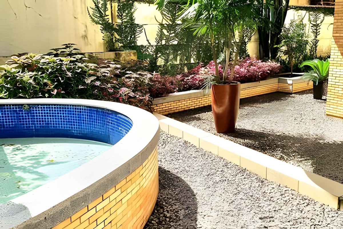An outdoor area features a curved pool with blue tiles, surrounded by a landscaped garden. Lush green plants and vibrant foliage are arranged along the path, offering a tranquil setting. A large, terracotta planter adds a warm touch to the space.