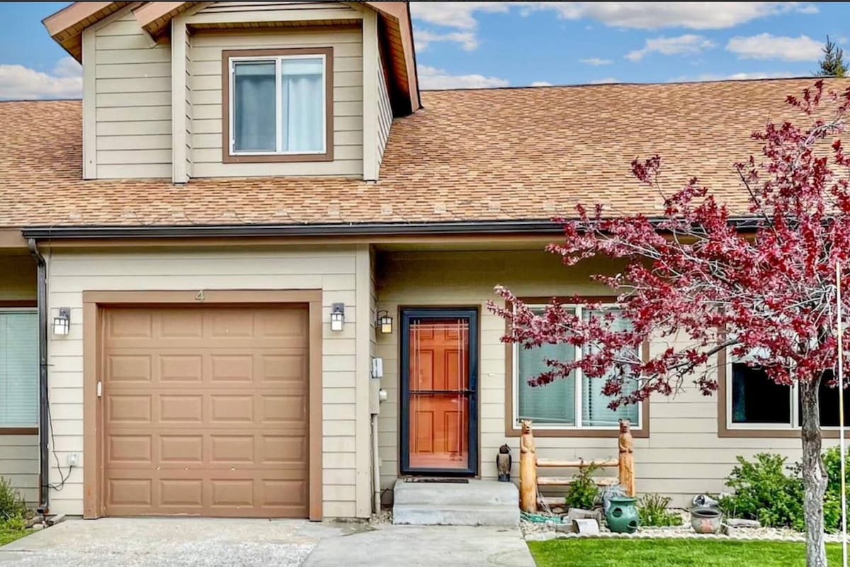 The exterior of the townhome features a well-maintained façade with a warm brown door and a garage. A blooming tree with red foliage is positioned in the foreground, adding a touch of nature. Two decorative dog statues are placed beside the entrance, enhancing the welcoming appearance.