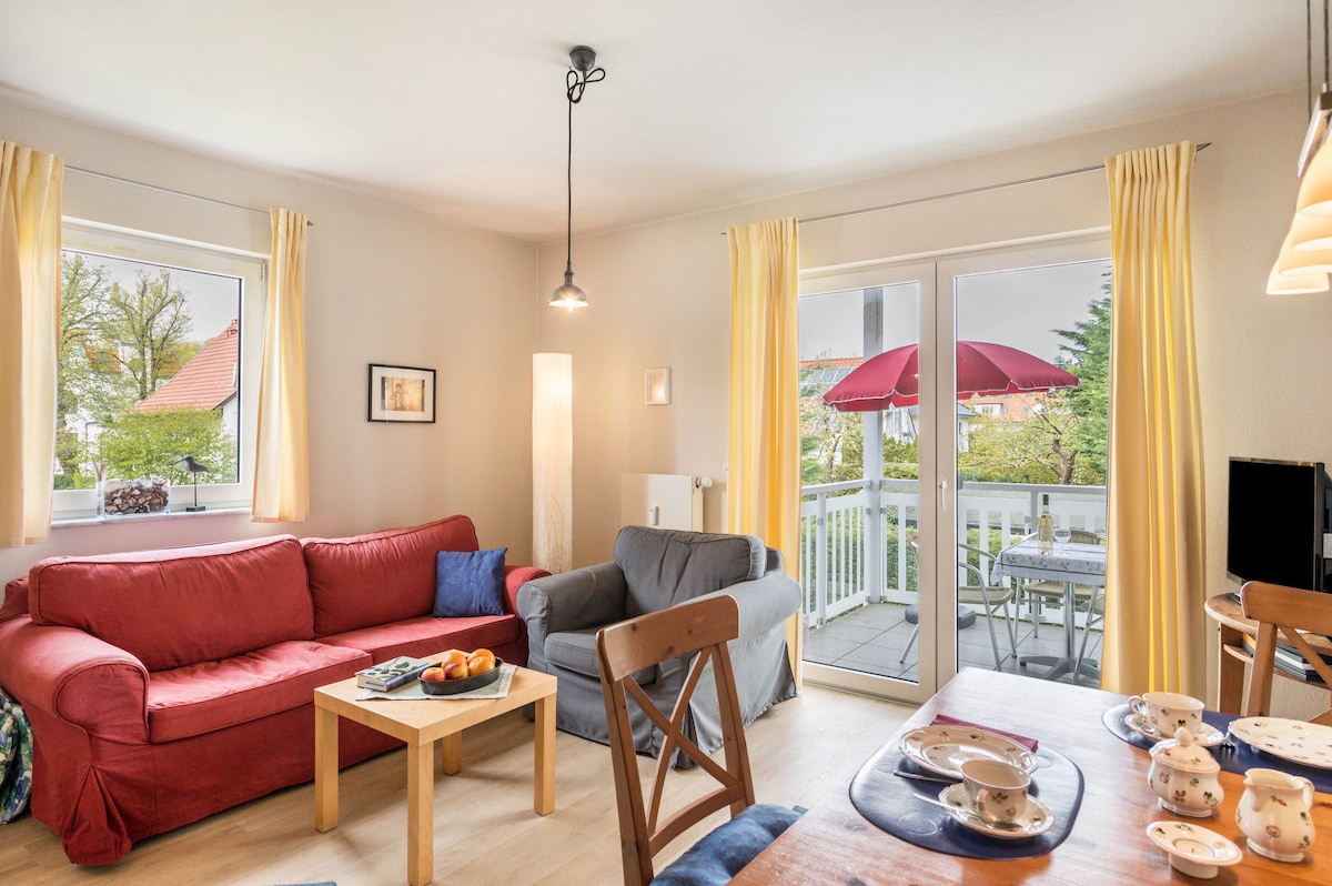 A living room is featured with comfortable seating, including a red sofa and a gray armchair. A coffee table is positioned at the center, adorned with a fruit bowl. Natural light is invited through two windows, and a sliding glass door leads to a balcony with outdoor seating.